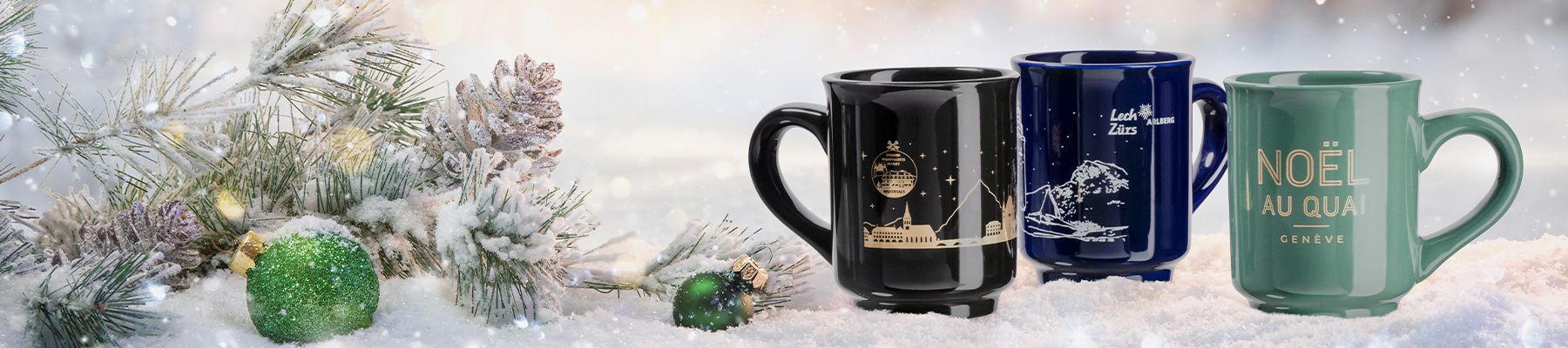 Drei festliche Tassen mit goldenen Motiven auf Schnee, daneben ein verschneiter Tannenzweig und ein grünes Ornament, die eine winterliche Urlaubsszene schaffen.