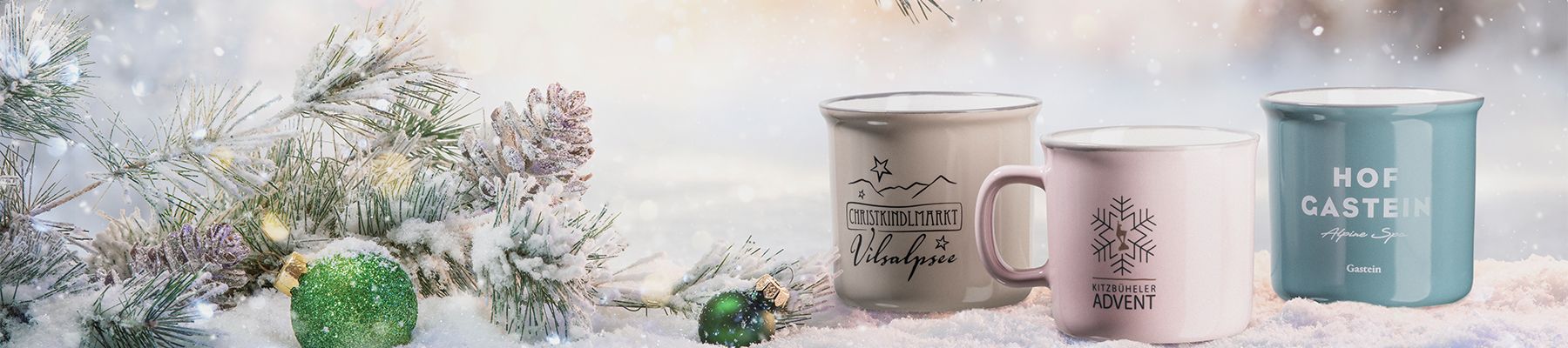 Drei festliche Tassen auf Schnee, jede mit winterlichen Motiven, daneben verschneite Tannenzweige und grüne Ornamente.