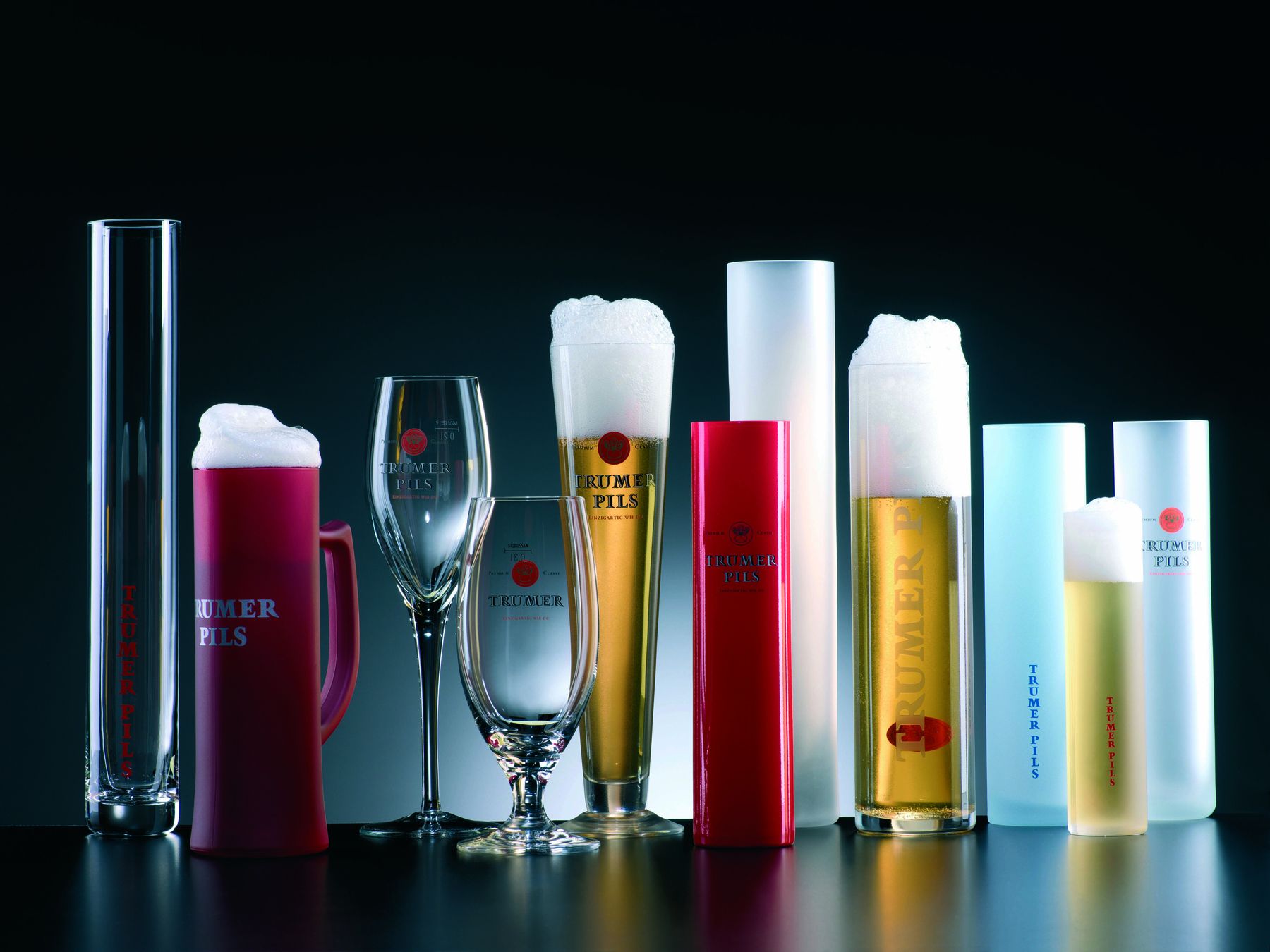 Eine Vielzahl von Trumer Pils-Gläsern und Krügen, jeweils gefüllt mit Bier und Schaum, angeordnet vor einem dunklen Hintergrund.