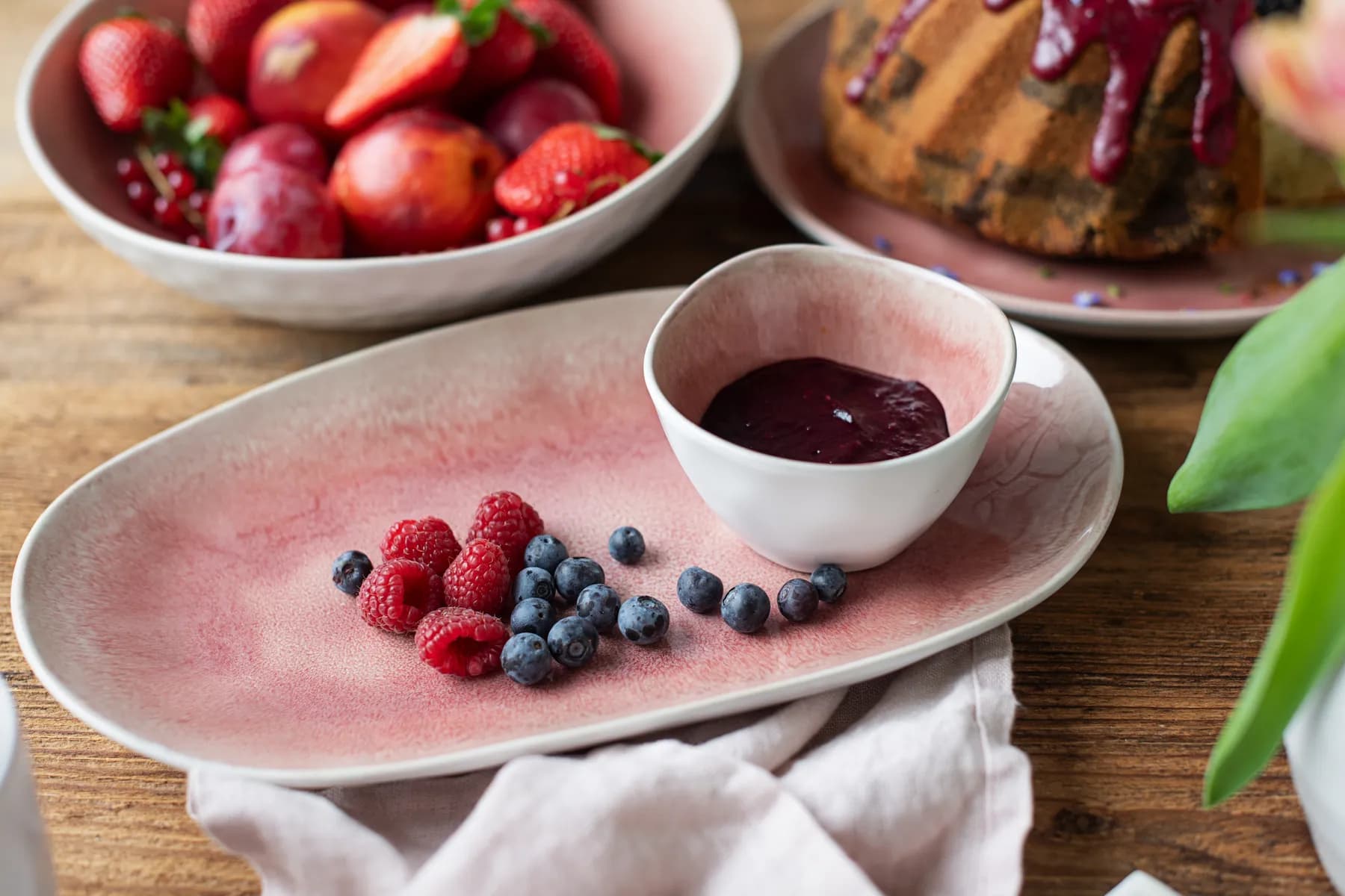 A pink plate with raspberries and blueberries, beside a cup of berry sauce. Background includes a bowl of fruit and a cake.